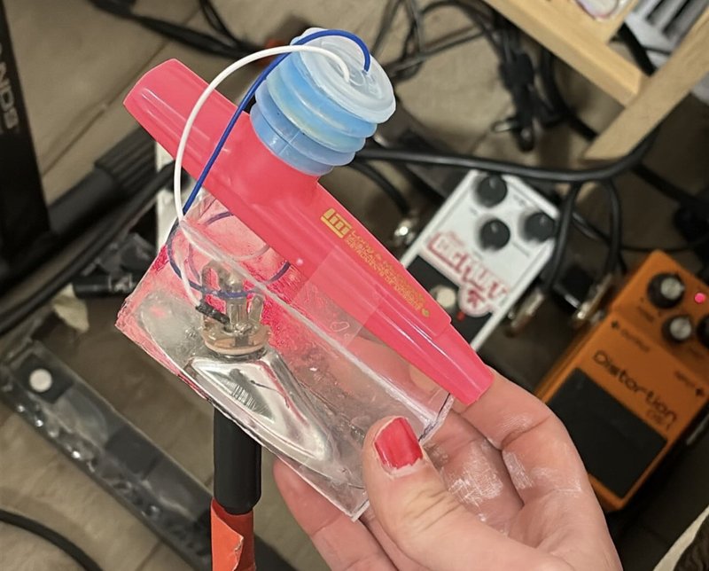 The finished electric kazoo held up in front of a pedalboard, with the blue resonator cap and clear CD-case cover