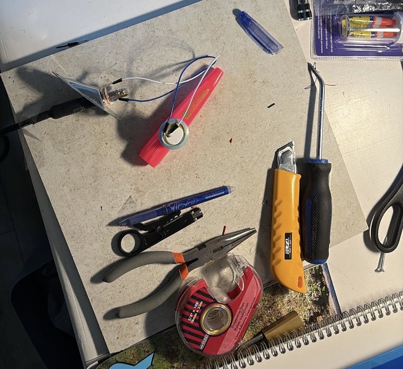 The kazoo on a workbench with its piezo disc and jack wired up, among pliers, a box cutter, a screwdriver and tape